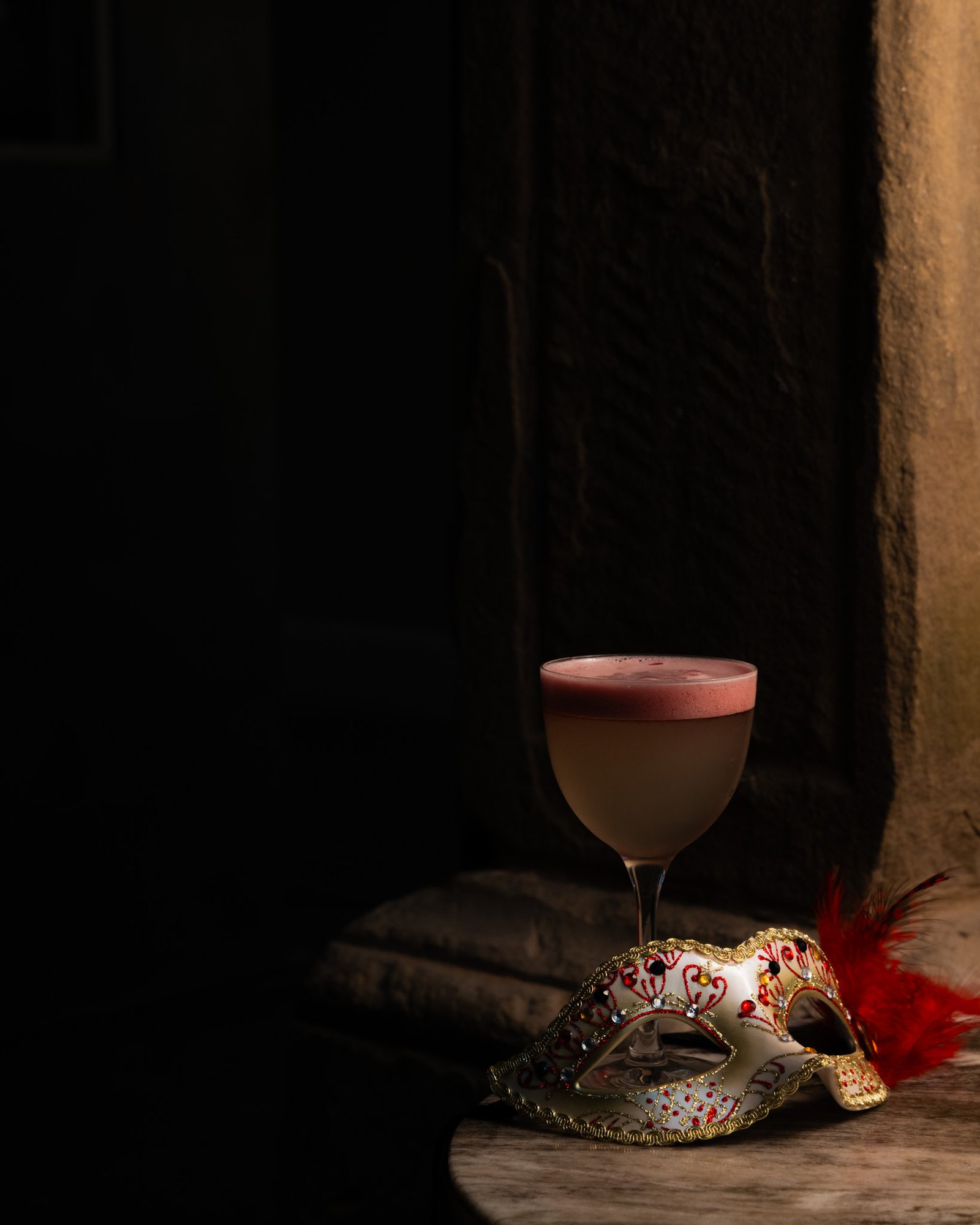 A feathered masquerade mask beside a pink-topped cocktail at Moods